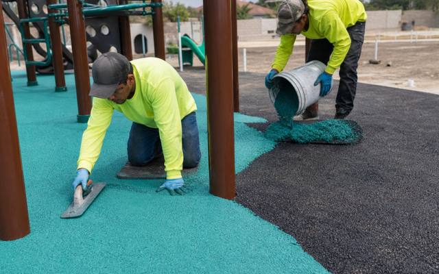 Two installers pouring teal rubber surfacing under a playground climber